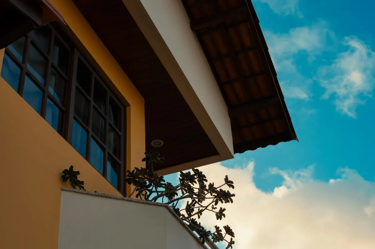 Fachada de casa com parede amarela, janelas de vidro escuro, beiral de telhado e céu azul com nuvens ao entardecer